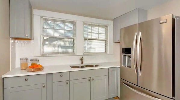 A modern kitchen with grey cabinets, a stainless steel refrigerator, a double sink, and a window with blinds.
