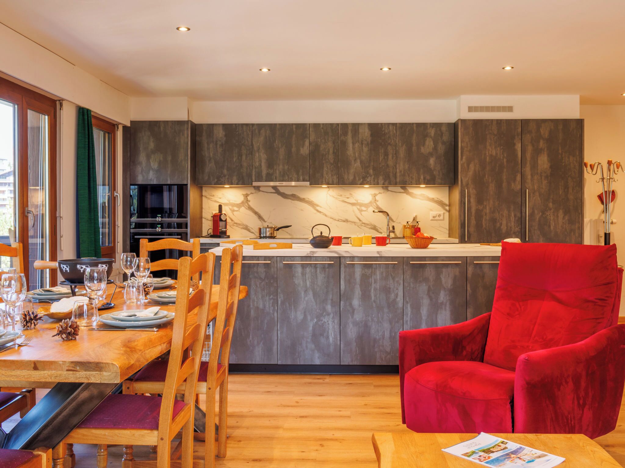 Une cuisine moderne avec des meubles de couleur sombre, un pare-feu en marbre et un coin repas avec des chaises en bois et un fauteuil rouge.