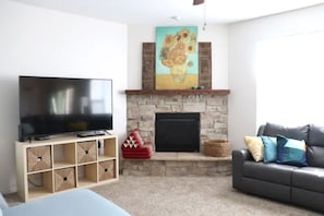 Uma sala de estar com televisão de tela plana em um suporte de madeira, lareira de pedra, sofá e um quadro sobre a lareira.