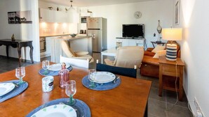 A dining area with a wooden table set for four, a kitchen in the background, and a living area with a sofa and a TV.