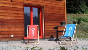 Un bâtiment en bois avec une porte rouge, deux fauteuils d’extérieur portant la marque « inter », et une petite table avec un pot de fleurs.