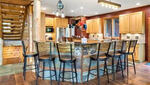 A modern kitchen with wooden cabinets, a central island with bar stools, hanging pots, and a chandelier.
