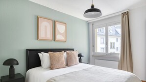 Un dormitorio ordenado con cama, mesitas de noche, ventana con cortinas y dos cuadros enmarcados en la pared.