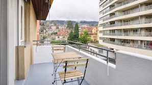 Un balcón con mesa y sillas de madera, con vista a un paisaje urbano que incluye edificios y una montaña en la distancia.