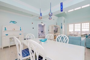 A room with a dining table, chairs, a sofa, and a TV. There are hanging lights and a view through a window.