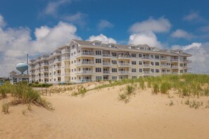 Un hôtel à plusieurs étages, doté de balcons, situé sur une plage de sable fin bordée de dunes.