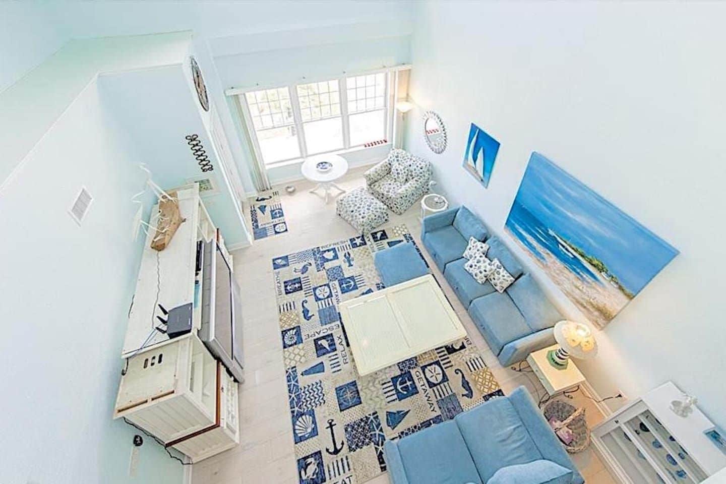 Una sala de estar con un alfombra estampado, un sofá azul, un cuadro de una playa y una ventana con persianas.