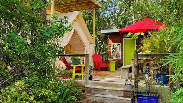 Une terrasse en bois mène à une petite maison surmontée d’un parasol rouge, le tout étant entouré d’une végétation luxuriante.