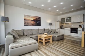 Ein modernes Wohnzimmer mit einer Eckcouch, einem Couchtisch aus Holz und einer Küchenzeile mit weißen Schränken und Geräten.