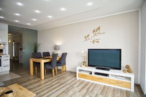 A modern living room with a dining area, a flat-screen TV on a white console, and a wall-mounted decorative piece. - Vacation apartment near the harbor with sea view - Hafenresidenz Lauterbach FeWo 2-2 - (Putbus)