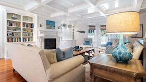 A spacious living room with a beige sofa, a wooden coffee table, and a blue vase. There is a fireplace, a bookshelf, and a large window with a view of greenery. - Center Of It All: 454-Cv  Sea Captain's Estate with Heated Pool and Pickle Ball Court (Centerville)