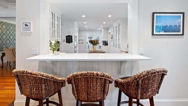 A kitchen with a central island, three wicker chairs, white cabinetry, and a framed picture on the wall. - Center Of It All: 454-Cv  Sea Captain's Estate with Heated Pool and Pickle Ball Court (Centerville)