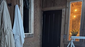 Interior de una cabaña de madera con una puerta negra, una toalla colgada y un cuenco metálico sobre un felpudo.