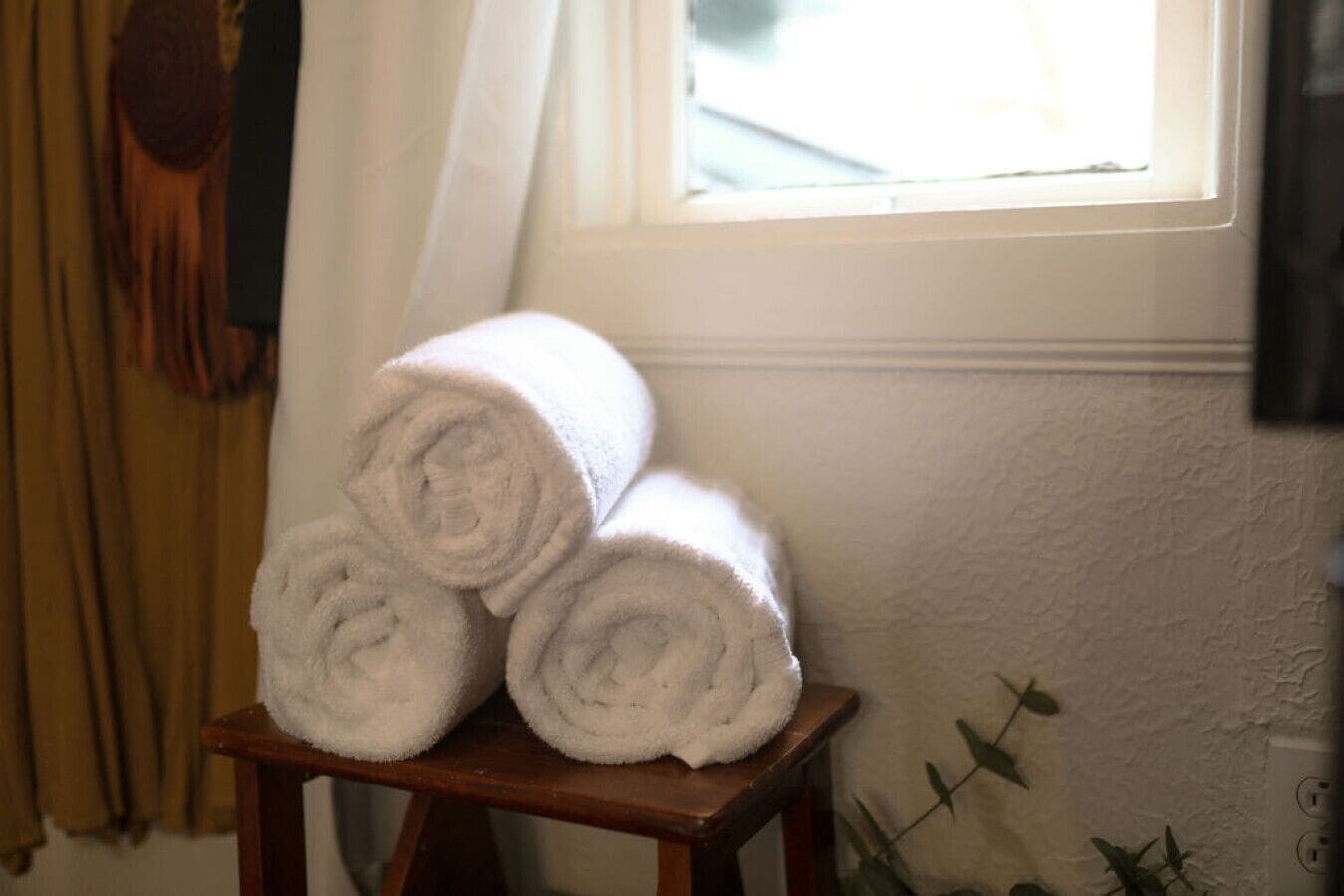 A stack of white towels on a wooden stool near a window with a curtain.