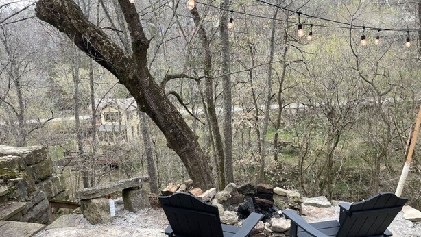 Dos sillas Adirondack negras mirando un brasero de piedra con una pared de piedra y un árbol al fondo.