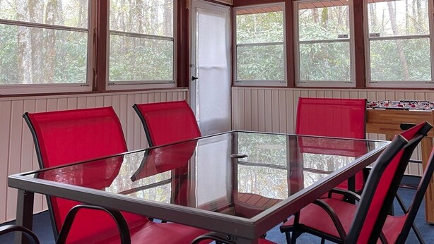 Une pièce avec une table à plateau de verre entourée de chaises rouges, des poutres en bois au plafond et de grandes fenêtres donnant sur la forêt.