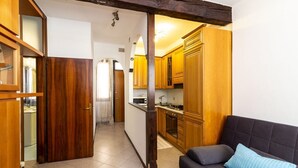 Una cuina petita amb armaris de fusta, forn de pared i un sofà fosc amb un coixí blau.