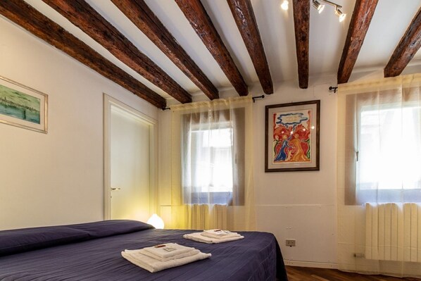 Un dormitorio con cama, dos toallas, vigas de madera, una ventana con cortinas y un cuadro en la pared.