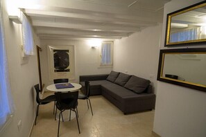 Un espacio habitable pequeño con un sofá gris, una mesa redonda con sillas y un cuadro en la pared.