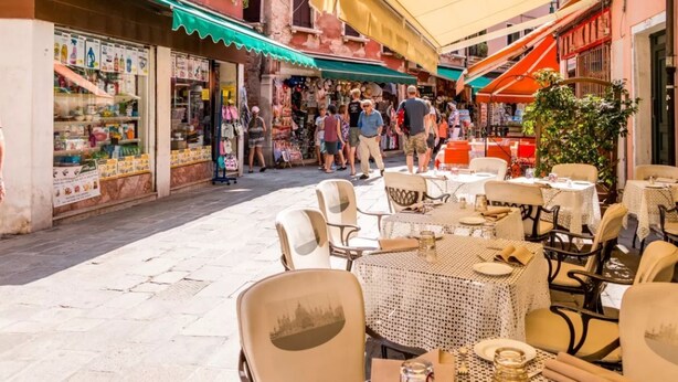Un café al aire libre con mesas y sillas, rodeado de tiendas y una calle estrecha.