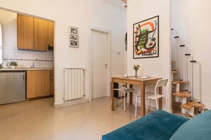 Uma cozinha compacta com armários de madeira, bancada branca e forno embutido. Possui uma área de refeições com mesa e cadeiras de madeira, e um sofá azul.