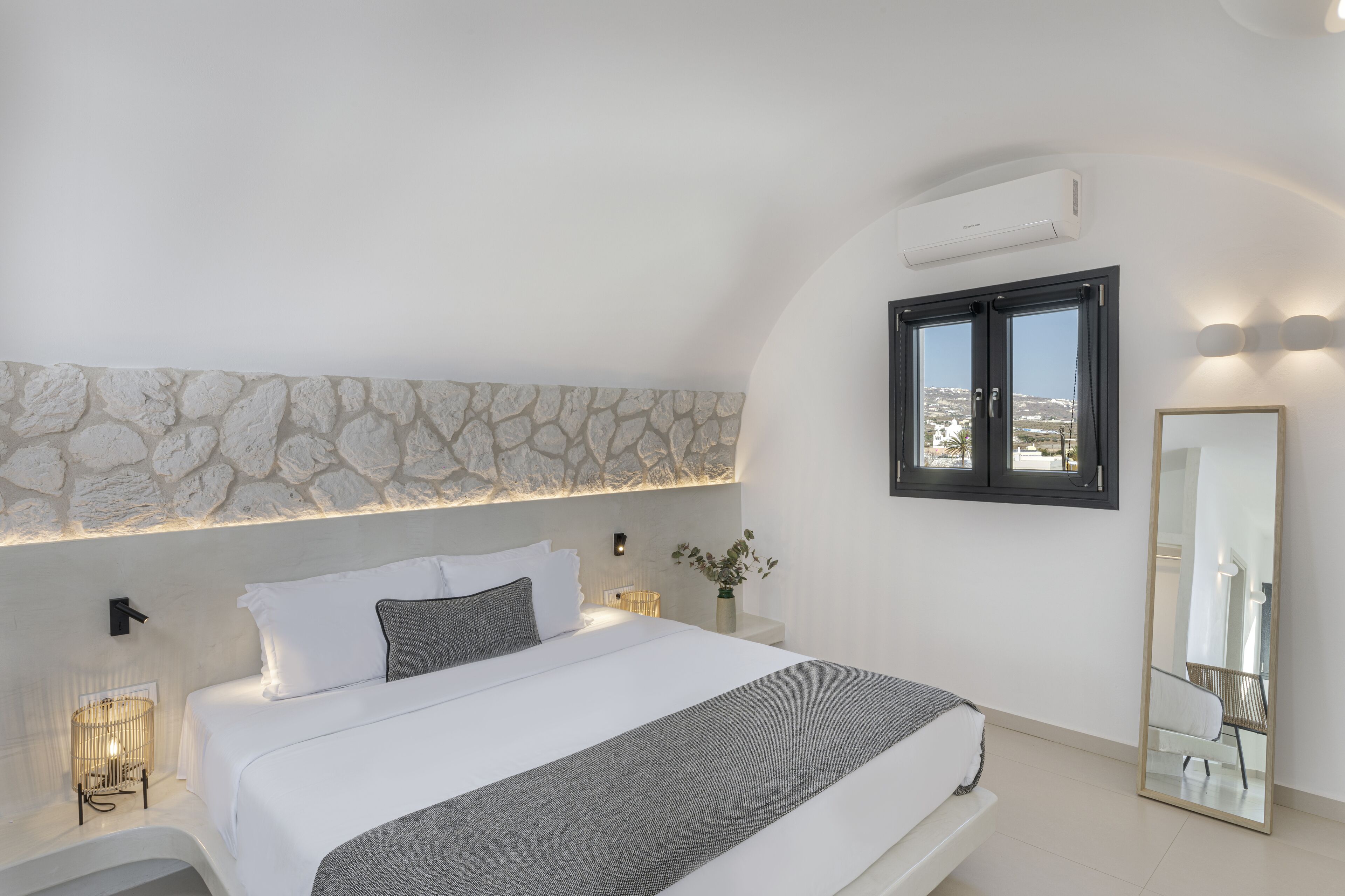 A modern bedroom with a stone accent wall, a large bed, bedside lamps, a mirror, and a view through a window.