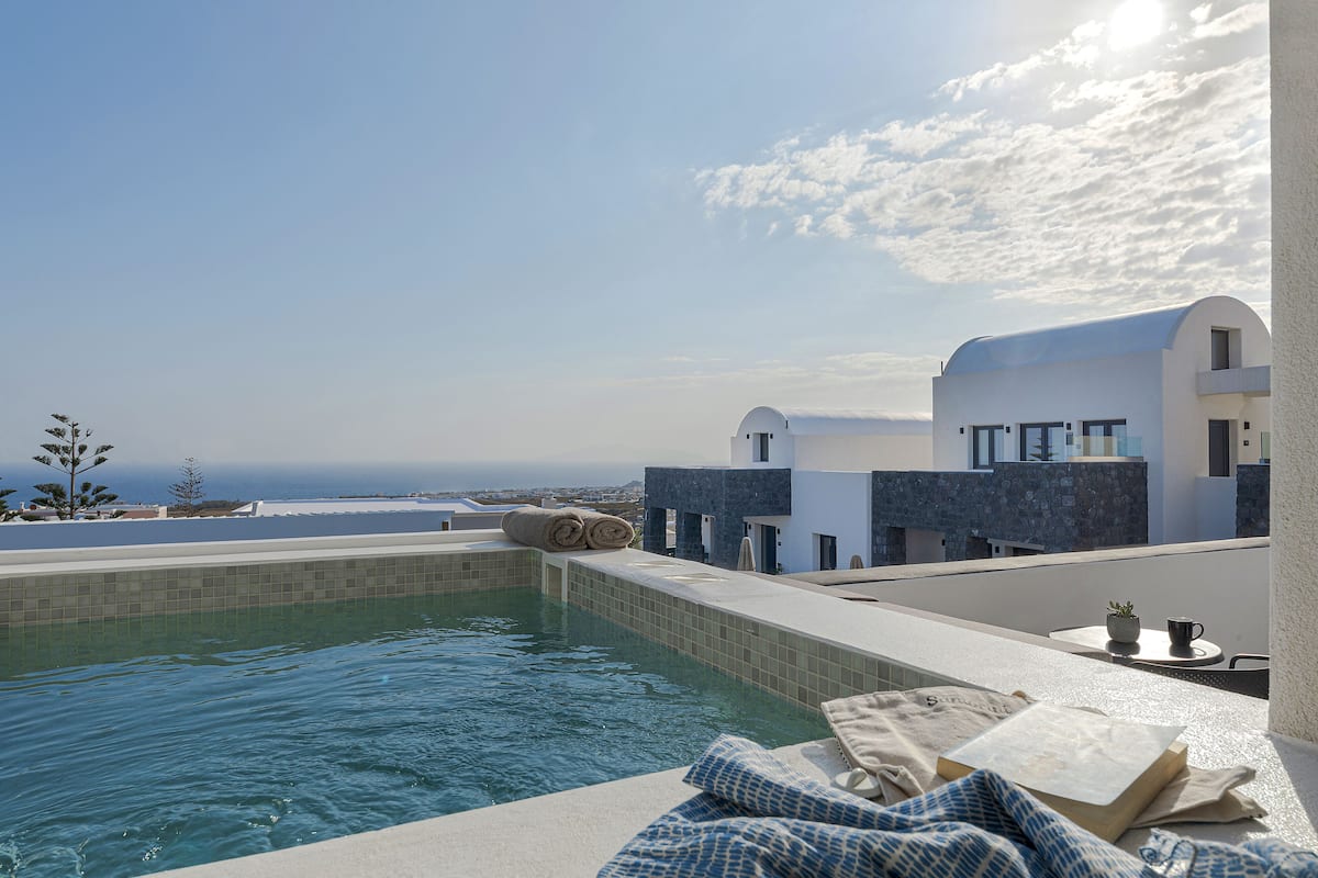 Une piscine à débordement située sur le toit, offrant une vue sur l’océan, un bâtiment blanc doté d’un toit incurvé, et une petite table sur laquelle se trouvent une plante et une tasse.