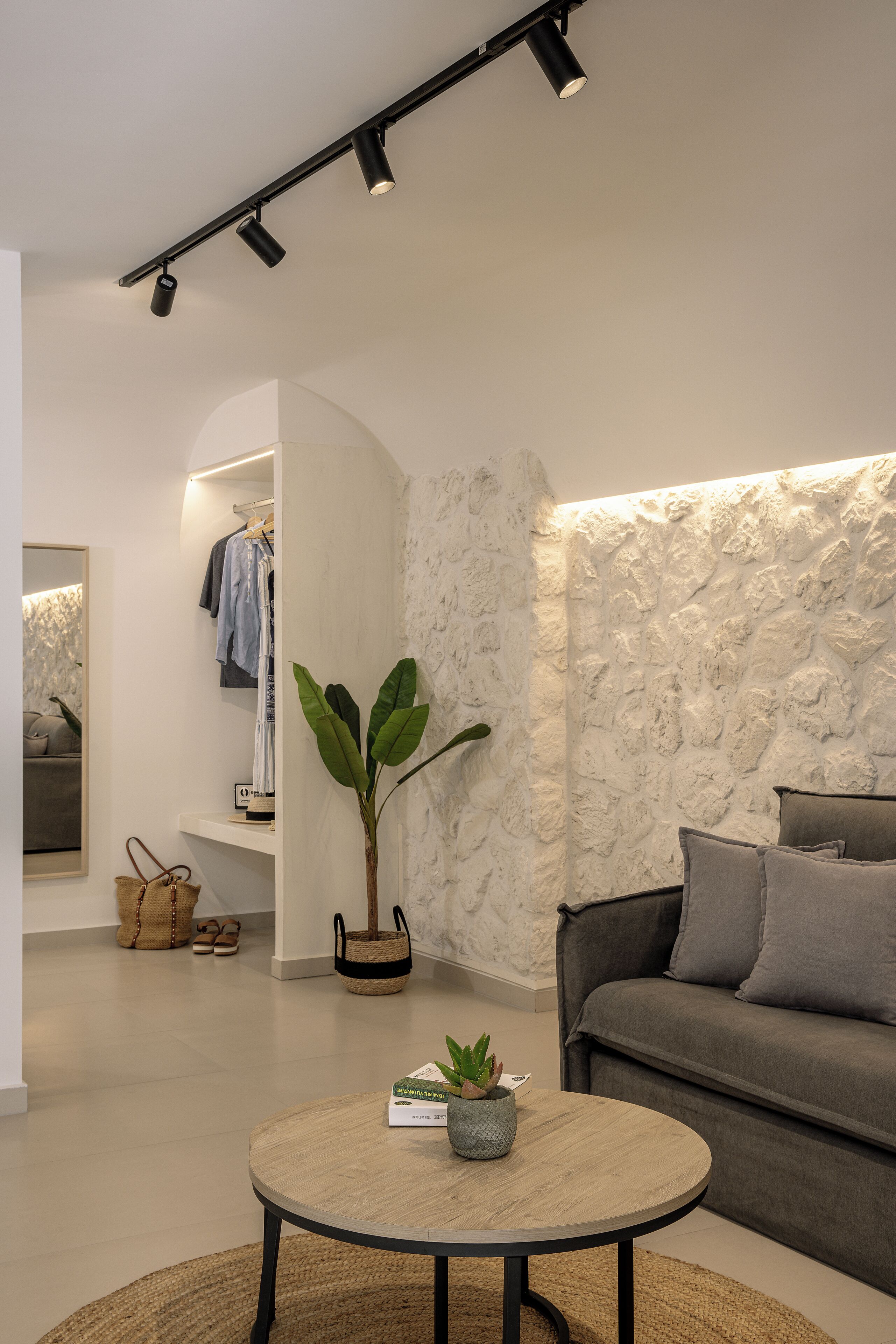 A modern living room with a round coffee table, a grey sofa, a stone-textured wall, and a hallway with a clothing rack and a mirror.