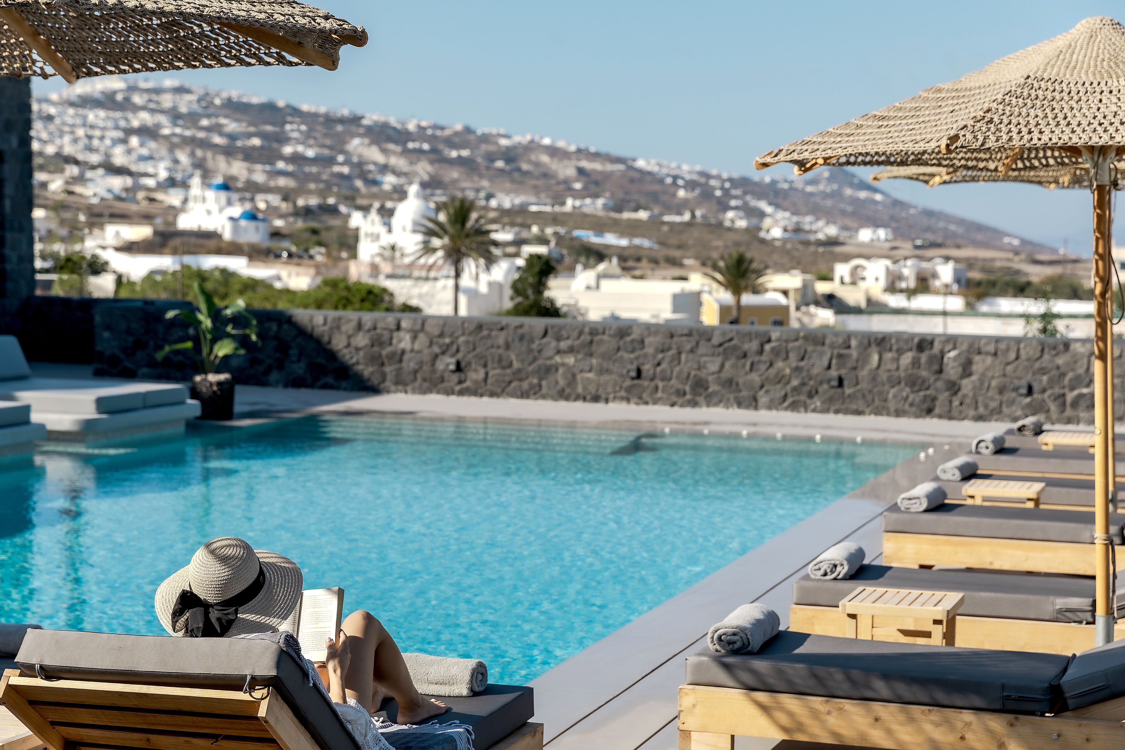 Un área junto a la piscina con sillones de descanso y sombrillas, una pared de piedra y vistas a una colina con edificios.