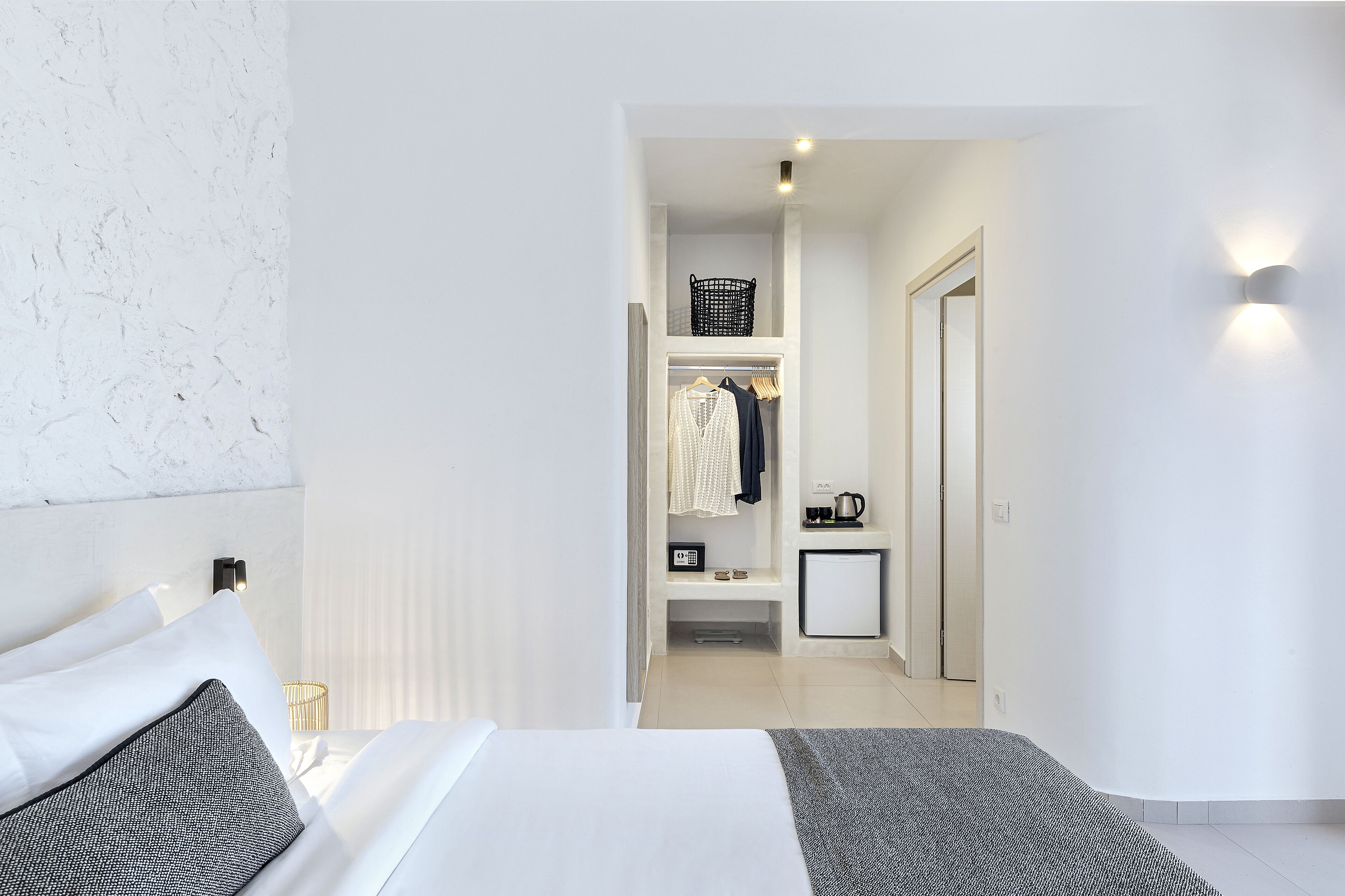 Una habitación de hotel moderna con una cama bien hecha, una mesita de noche pequeña con lámpara, un armario con ropa colgada y una kitchenette compacta.