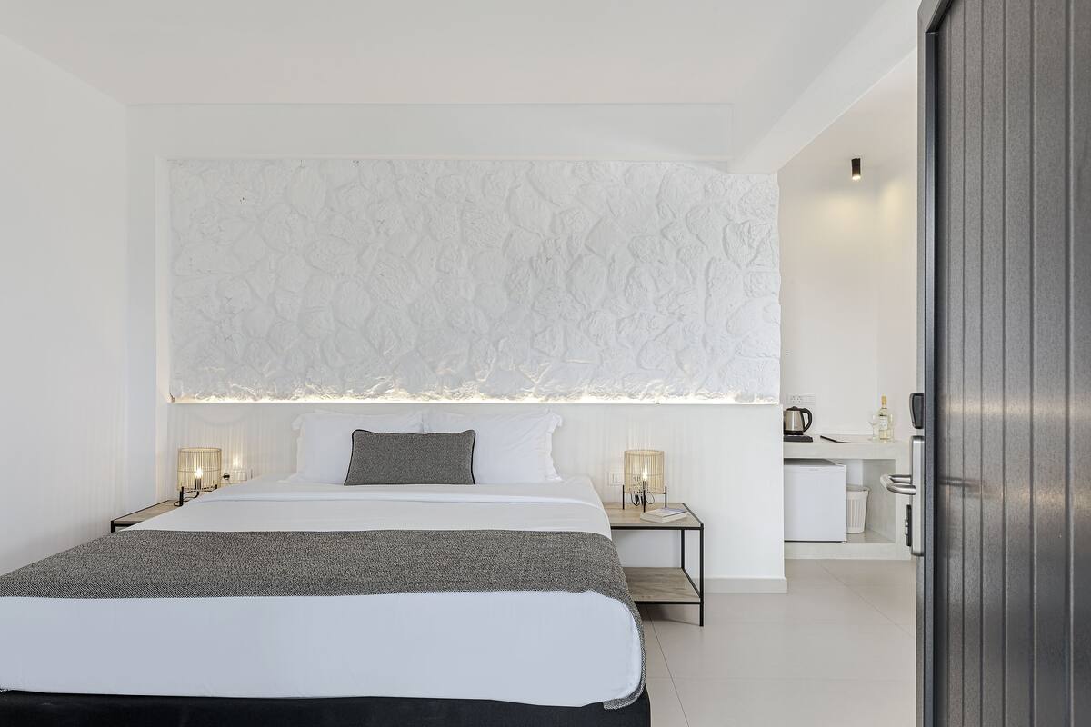 Une chambre d’hôtel moderne avec un grand lit, des tables de chevet, une petite kitchenette et un mur blanc à texture.