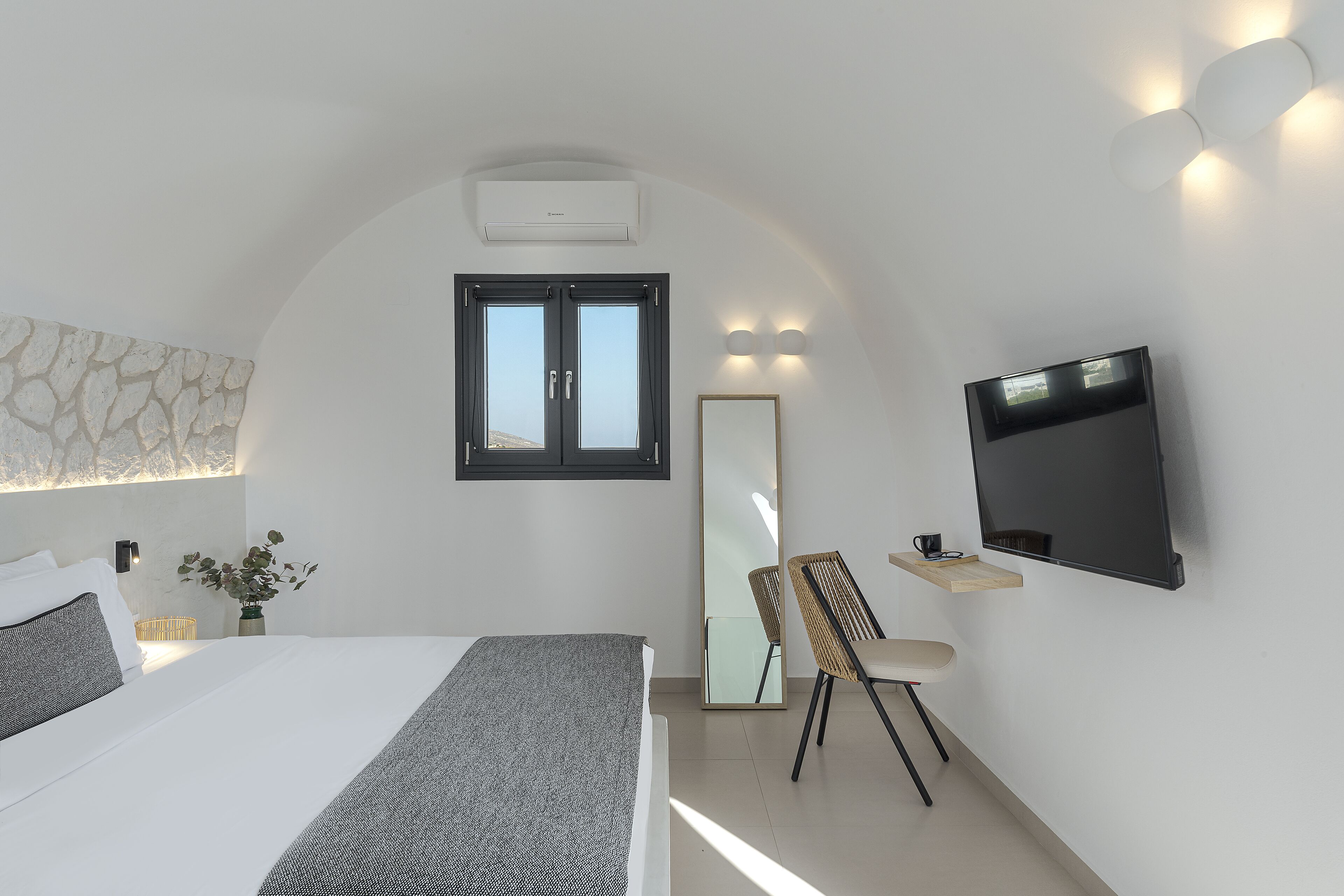 A modern bedroom with a large bed, a flat-screen TV, a chair, a small table, and a wall-mounted mirror.