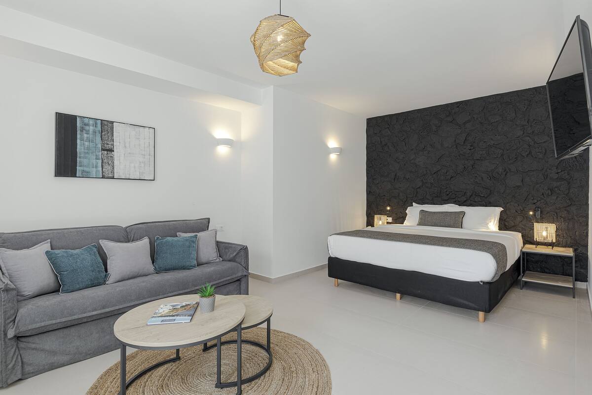 Une chambre moderne avec un canapé gris, une table basse ronde, un lit avec une tête de lit noire et un téléviseur fixé au mur.