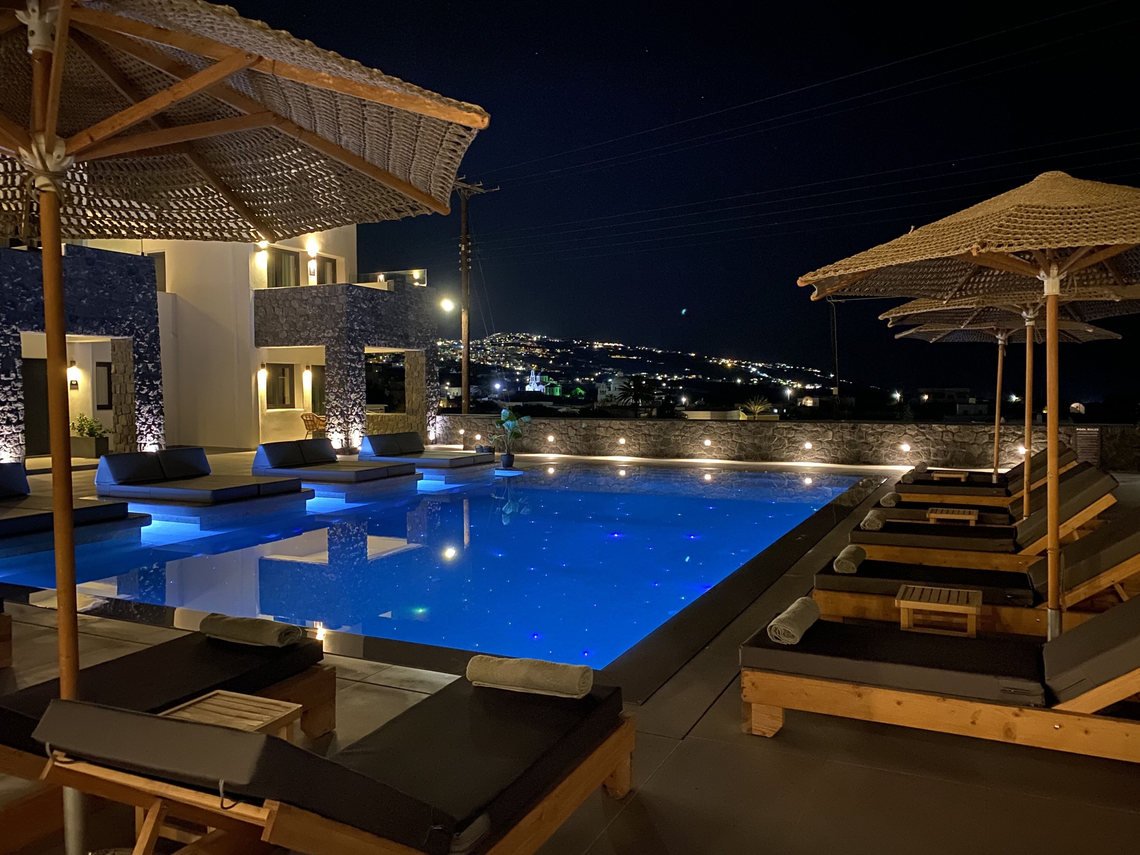 Ein beleuchteter Poolbereich bei Nacht mit Liegestühlen und Sonnenschirmen, ein Steinhaus und ein beleuchteter Hügel im Hintergrund.