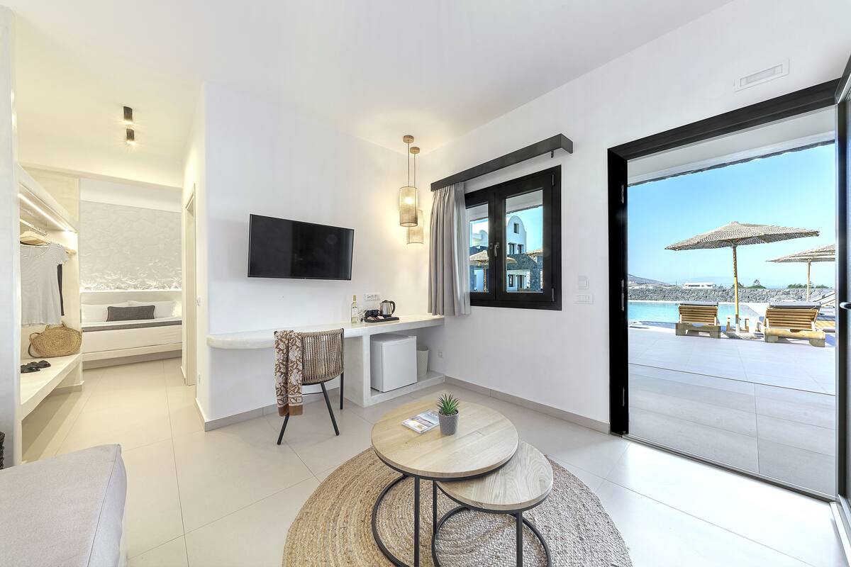 Une chambre moderne et bien éclairée, dotée d’un téléviseur à écran plat, d’un petit bureau et d’un coin salon confortable avec une table ronde.