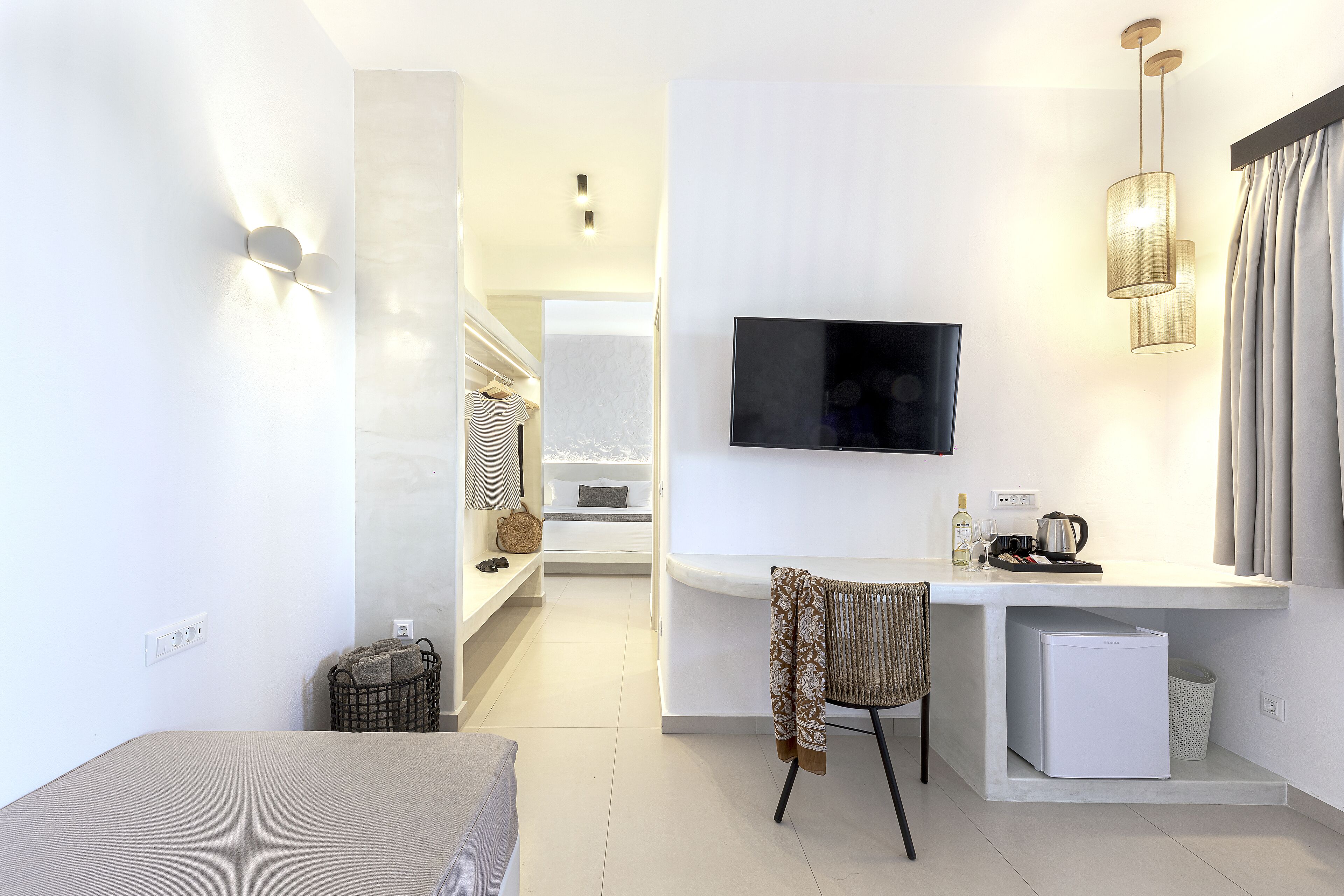 A modern hotel room with a bed, a desk with a television, a chair, and a small refrigerator.