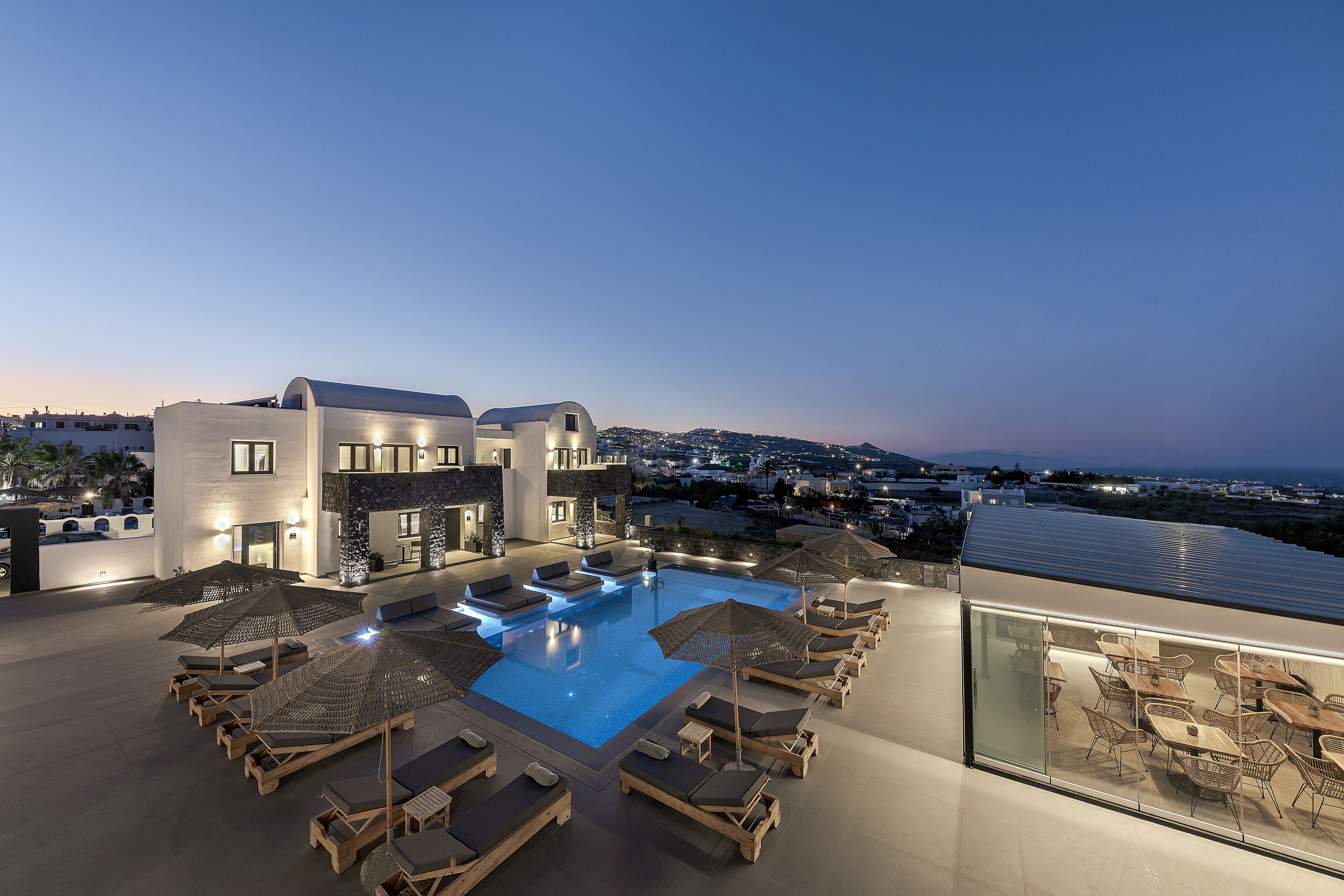 A rooftop pool area with sun loungers and umbrellas, a pool, and a view of the city at dusk.