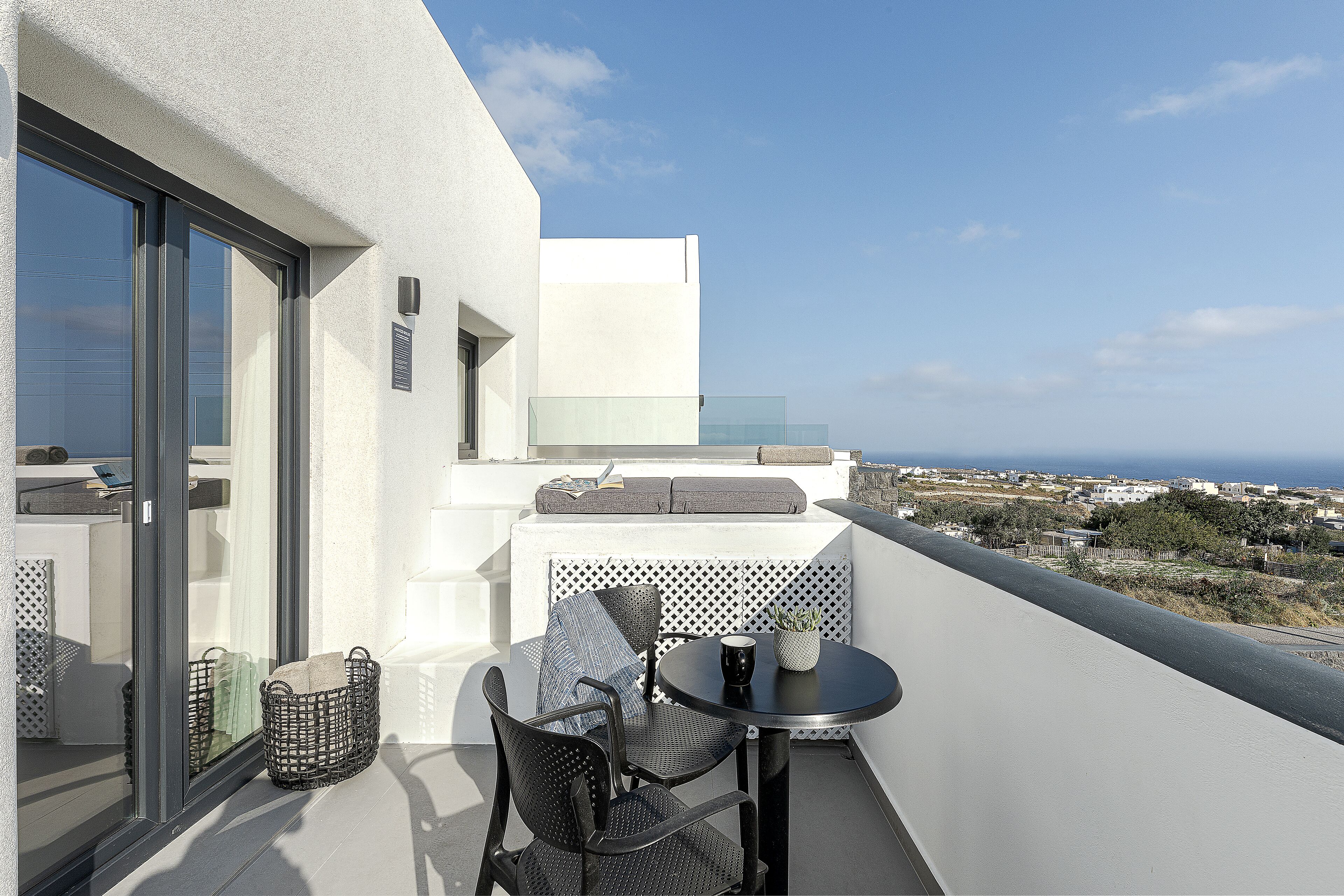 A balcony with a table and chairs, a wicker basket, and a view of the sea and buildings.