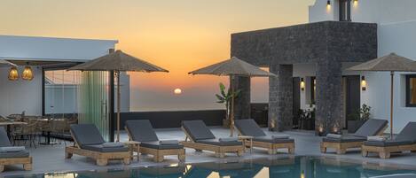A poolside area with lounge chairs and umbrellas, a dining area, and a view of the sunset.