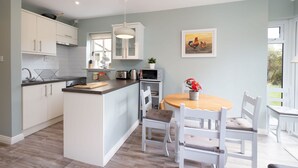 A modern kitchen with white cabinetry, a central island, and a dining area with white chairs and a wooden table. - Cottage 420 - Clifden - sleeps 4 guests  in 2 bedrooms (Clifden)