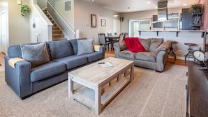 A modern living room with a grey sofa, a wooden coffee table, and a red throw blanket. A staircase leads to another level, and a kitchen area is visible in the background. - Meritage: European-inspired Family Home (Corvallis)