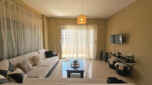 A living room with a sofa, a coffee table, a TV, and a large window with curtains. - Albania Dream Holidays Accommodation (Sarand)