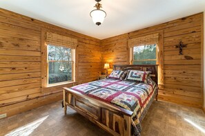A wooden cabin bedroom with a bed, windows, and a cross on the wall.