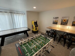 A room with a foosball table, a table tennis table, and a foosball table with a 'Goal' logo. There are two chairs and a small table with a lamp. - Most Spacious 2075 sq ft Paradise Townhome near Lake in heart of Garden City (Garden City)
