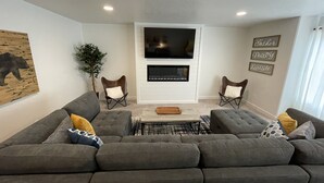 Ein Wohnzimmer mit einem grauen Ecksofa, zwei Sesseln, einem Flachbildfernseher, einem Kamin und einer Wand mit motivierenden Zitaten.