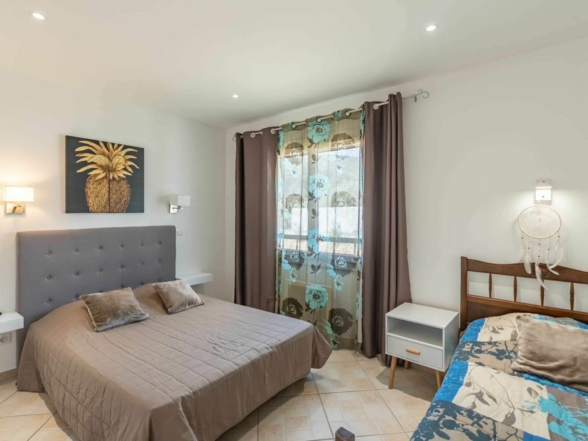 Um quarto com uma cama grande, uma mesa de cabeceira, um aparador, uma janela com cortinas, um abajur fixado na parede e um sonho apanhador.