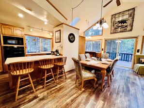 A wooden dining table with chairs, a kitchen area with a microwave, and a large window overlooking a forest.