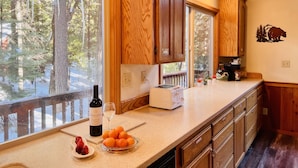 Una cocina con armarios de madera, un ventanal con vistas a árboles, una botella de vino y un cuenco de naranjas.