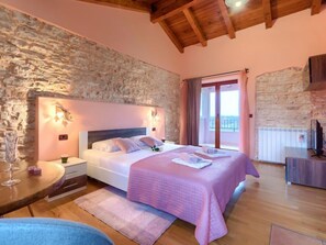 Quarto com parede de pedra, teto de madeira, cama com colcha rosa, mesa de cabeceira e vista para o exterior.