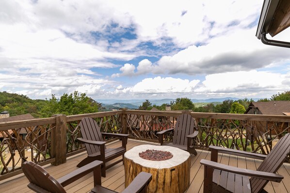 A wooden deck with Adirondack chairs and a central fire pit, overlooking a mountainous landscape.